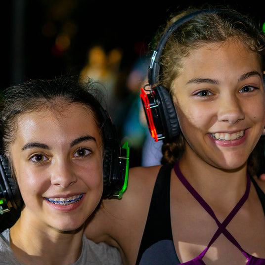 Quiet Events Silent Disco headphones for kids. A pair of girls side to side embracing and having fun with headphones.