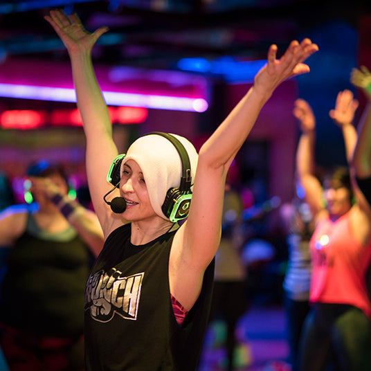 Quiet Events Silent Disco headphones for fitness and meditation. A fitness instructor providing a class to her students with her hands up and wearing headphones.