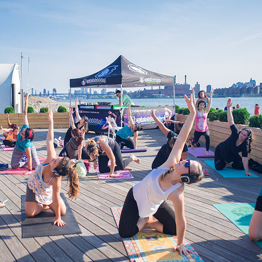 Quiet Events Silent Disco headphones for fitness and wellness. A yoga instructor with her students outdoors wearing headphones and posing.