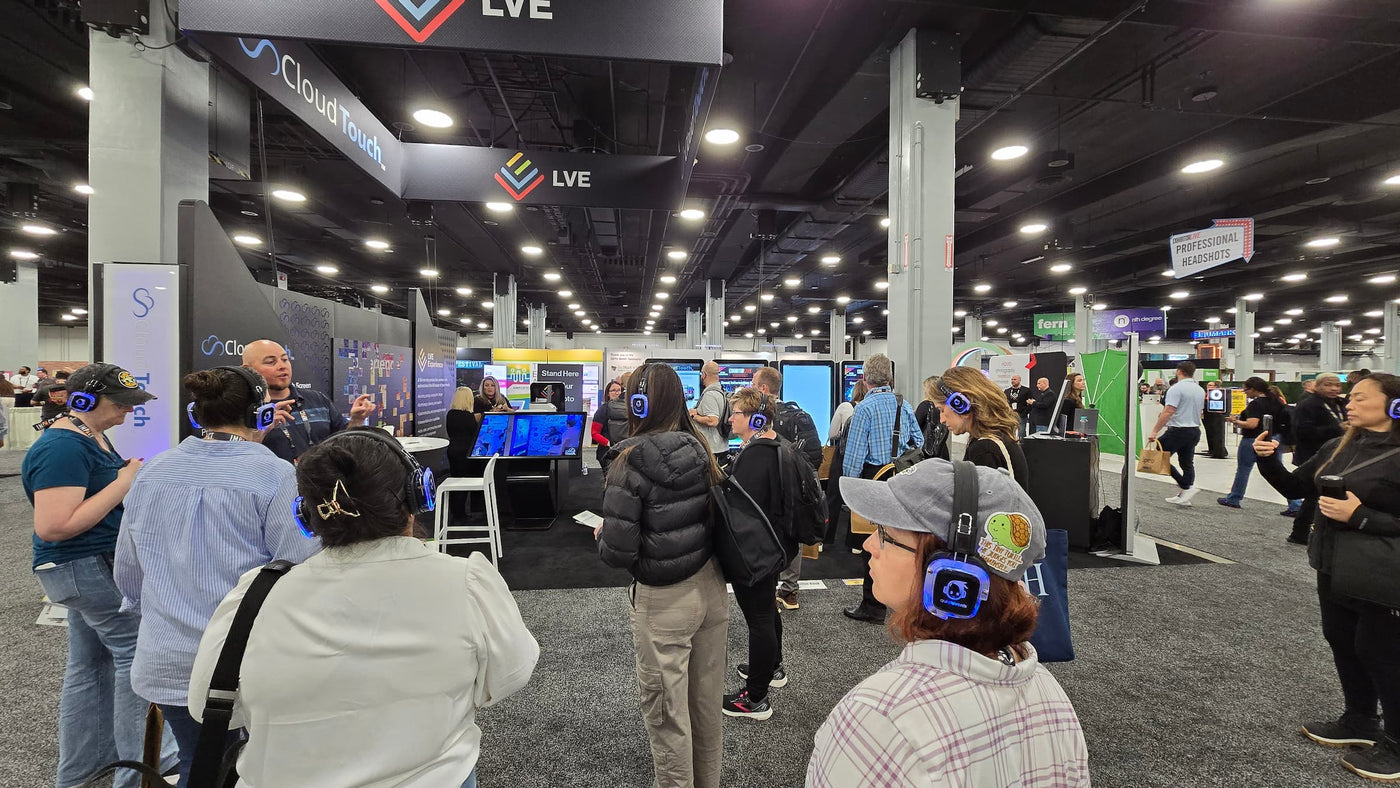 Quiet Events Silent Disco headphones for conventions a group of people standing with headphones listening to a speaker at a booth.