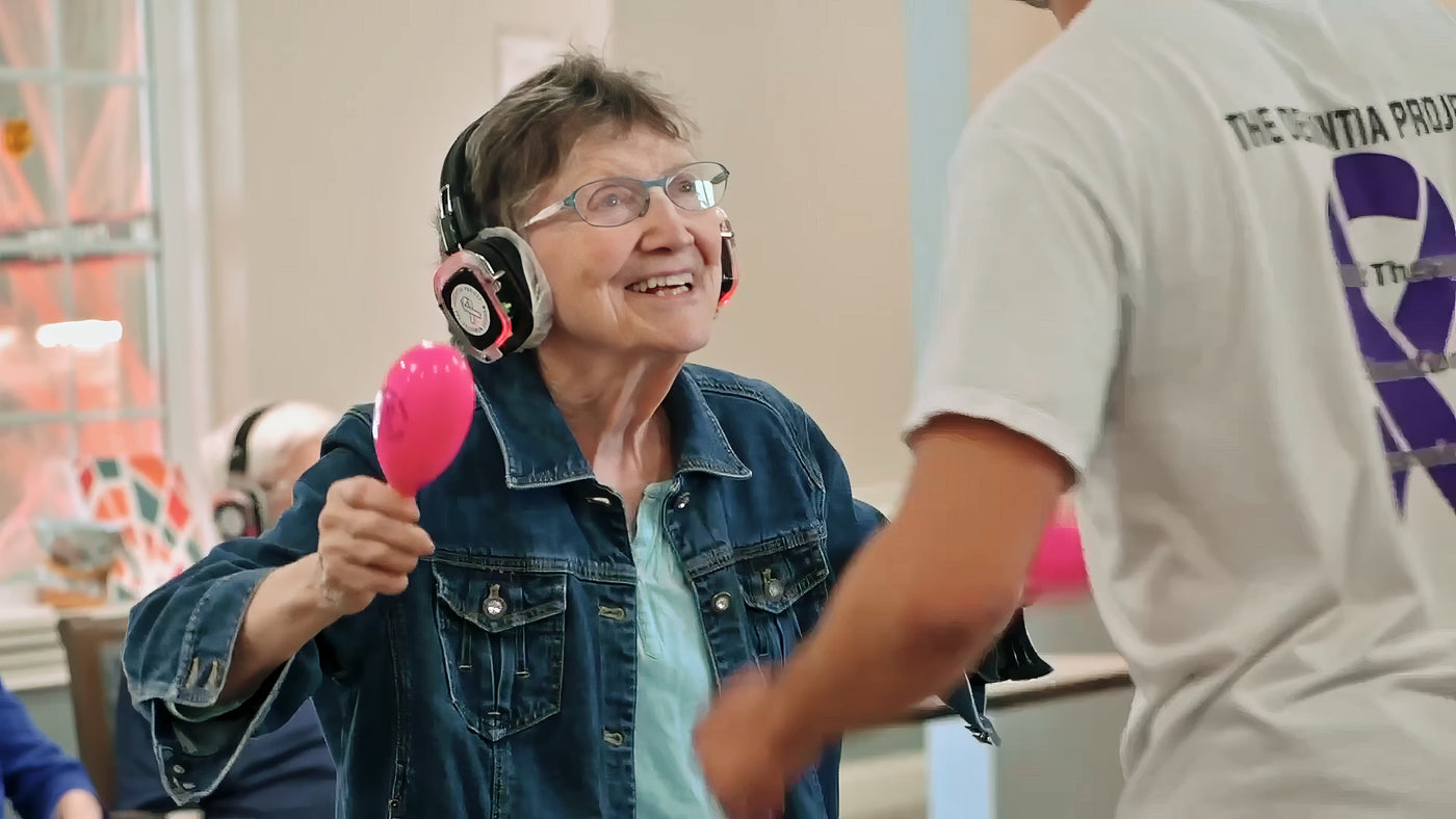 Quiet Events Silent Disco headphones for senior care. A older woman having fun being entertained holding maracas and wearing headphones.