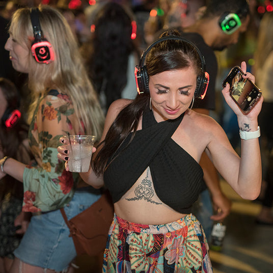 Quiet Events Silent Disco headphones for traveling. A young woman dancing and having fun outdoors with others wearing headphones.