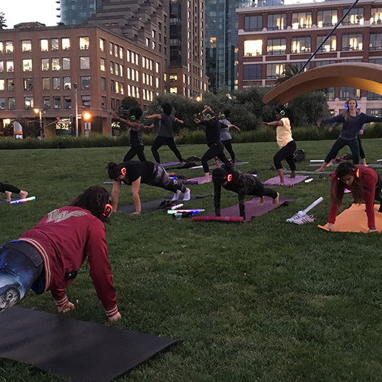 Quiet Events Silent Disco headphones for yoga. A group of people wearing headphones at a park doing yoga poses.