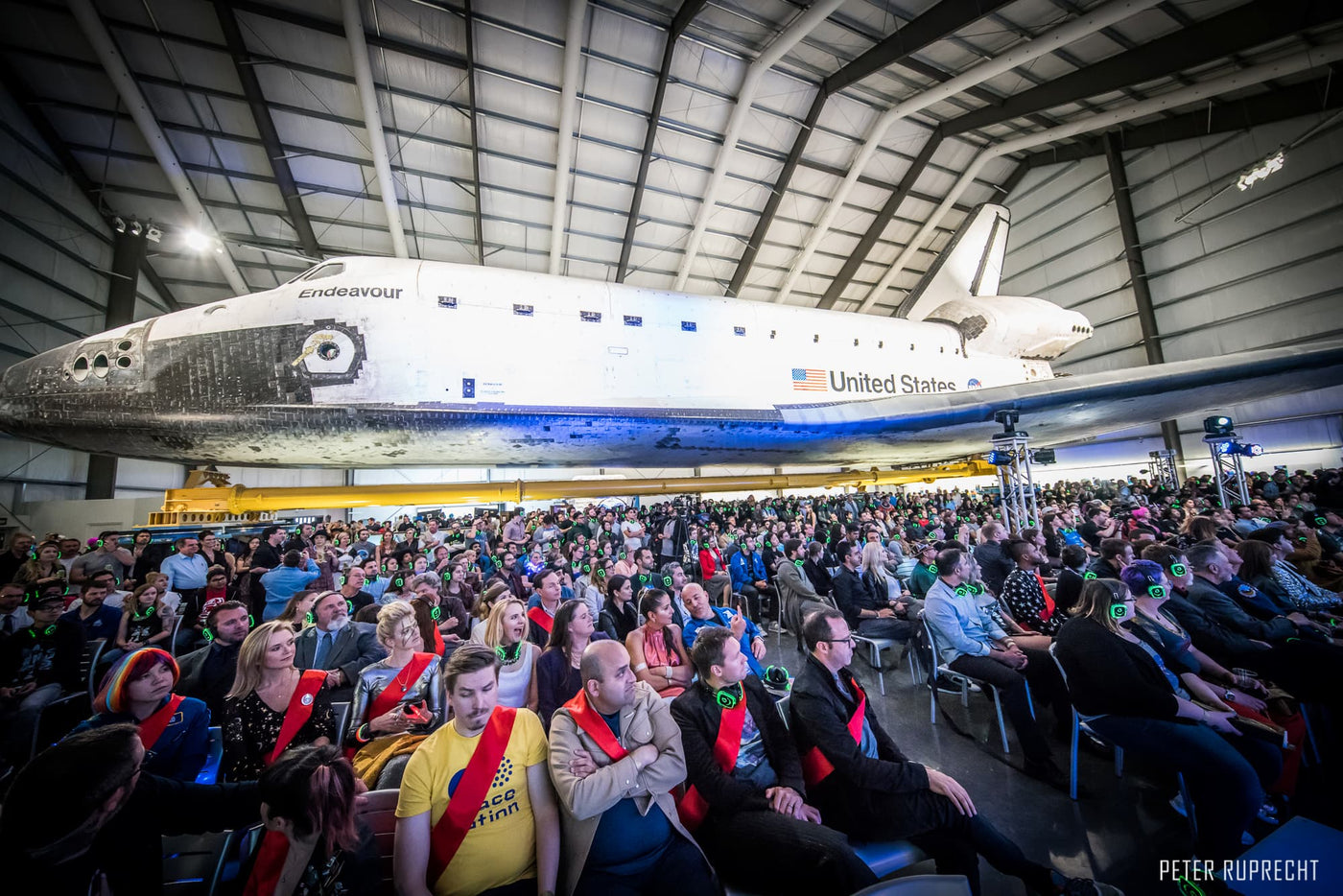 Silent Disco headphones for other events. A group of people gather and listening carefully during a presentation with a NASA space shuttle suspend in the air wearing headphones.